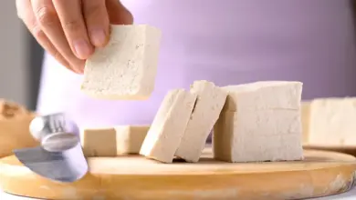 Ruce drží kus čerstvého tofu nad dřevěnou prkénkem s nožem.