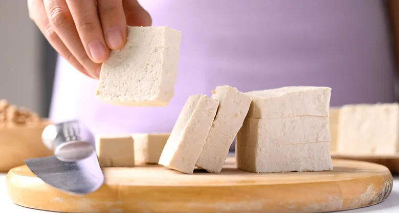 Ruce drží kus čerstvého tofu nad dřevěnou prkénkem s nožem.