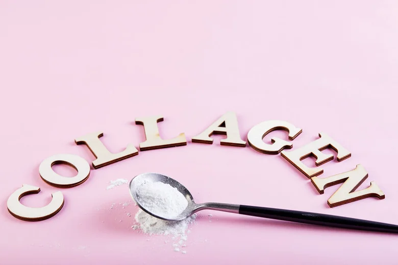 Lžíce bílého kolagenu před dřevěnými písmeny COLLAGEN na růžovém pozadí.